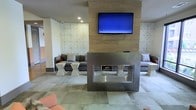 This single-floor space presents a sleek, modern lounge area designed with a refined aesthetic. The focal point is a striking double-sided stainless steel fireplace, which serves as a central divider, creating distinct yet connected seating zones on either side. Mounted directly above on a warm wood-paneled accent wall is a large flat-screen television, adding a media-forward element to the space. The surrounding walls feature an eye-catching geometric tile pattern with an embossed star or sunburst design in a neutral tone, contributing texture and visual interest without overwhelming the overall palette. Seating areas are furnished with low-profile gray sofas and modern hourglass-shaped side tables in white, complemented by occasional accent chairs in a muted coral tone. The flooring consists of large-format tiles in varying neutral shades, laid in a patchwork pattern that adds subtle dimension underfoot. Recessed lighting illuminates the space evenly, enhancing the clean lines and contemporary finishes throughout. The overall ambiance is polished and inviting, suited for social gathering or relaxed lounging.