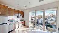 This single-floor space offers a clean, modern aesthetic defined by crisp white walls, light hardwood-style flooring, and an abundance of natural light. The kitchen is a standout feature, equipped with rich dark-wood cabinetry that extends to the ceiling for generous storage, a full suite of stainless steel appliances including a refrigerator, dishwasher, range, and built-in microwave hood. Light countertops and a functional sink complete the well-organized layout. A striking floor-to-ceiling sliding glass door and window unit floods the kitchen and adjoining area with sunlight while offering elevated views of the surrounding neighborhood rooftops and streetscape. The living area is an open, versatile space with the same warm flooring and neutral white walls, creating a calm and adaptable environment. The bathroom is tastefully finished with large-format light tile flooring, a wood-toned vanity with a white countertop, and a clean, bright layout that includes a tub and toilet. Recessed lighting throughout the space enhances its modern, polished feel. Overall, the unit presents a cohesive, contemporary design that balances functionality with understated style.