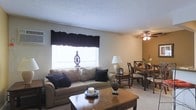 This single-floor space presents an open-concept layout that seamlessly connects the living and dining areas into one cohesive environment. The living area is anchored by a comfortable beige sofa adorned with navy accent pillows, positioned beneath a large window dressed with dark valance curtains that allow natural light to filter in. A warm-toned wooden coffee table sits at the center, complementing the neutral carpeted flooring that runs throughout the space. A table lamp provides additional ambient lighting, adding warmth to the room's overall atmosphere. Transitioning into the dining area, a wooden dining table is surrounded by classic cross-back chairs, with a ceiling fan overhead providing both light and airflow. Decorative touches, including a small floral arrangement and tabletop accents, add character to the dining space. The walls are painted in soft, neutral tones, and the textured ceiling contributes a traditional residential feel. Overall, the space offers a practical and welcoming layout suitable for everyday living and entertaining, with a cohesive design aesthetic maintained throughout both functional zones.