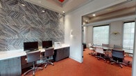 This single-floor business center is thoughtfully designed to support a variety of professional needs, combining focused workspaces with collaborative and casual areas. The entrance welcomes visitors with vibrant red accent walls, warm orange-red carpeting, and colorful modular seating, creating an energetic yet inviting atmosphere. Large windows and glass-paneled entry doors flood the space with natural light, offering views of the surrounding outdoor environment.

The dedicated computer workstation area features a built-in desk with multiple monitors, a multifunction printer, and dark wood cabinetry — all set against a dramatic floor-to-ceiling textured tile accent wall with a geometric chevron pattern, adding a striking design element to the room. Recessed lighting and wall sconces ensure a well-lit, functional workspace.

Adjacent to the workstation area, a private conference room provides a polished setting for meetings, furnished with a substantial wood conference table, ergonomic chairs, and two large windows fitted with sheer roller shades that diffuse natural light. A decorative wall sculpture adds an artistic touch.

A lounge nook rounds out the space, featuring comfortable upholstered chairs, small side tables, and bold abstract artwork displayed against the vivid red feature wall, making it ideal for informal gatherings or quiet relaxation.