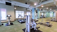 This fitness center offers a spacious, single-floor layout designed to accommodate a comprehensive workout experience. The open floor plan is anchored by high ceilings with exposed ductwork and piping, lending the space an industrial aesthetic that is both functional and modern. Structural columns painted in a soft blue-gray tone are distributed throughout, adding architectural character without interrupting the flow of the space.

Large windows along one wall invite abundant natural light, creating a bright and energizing environment for exercise. The flooring features a patterned green carpet interspersed with black rubber mats beneath the equipment, providing both comfort and safety.

The cardio section is prominently equipped with multiple treadmills and elliptical machines, positioned to take advantage of the natural light from the windows. The strength training area features a variety of cable and resistance machines, offering users a full range of exercise options. A large mirror spans a section of the wall, enhancing the sense of space and allowing users to monitor their form during workouts.

Overhead pendant lighting and recessed fixtures supplement the natural light, ensuring the space remains well-lit throughout the day. The overall ambiance is clean, open, and purposefully arranged to support an effective and comfortable fitness routine.