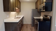 The Twain's 1-bedroom, 1-bathroom floor plan is designed for functional, comfortable living across a single level. The galley-style kitchen is a standout feature, showcasing rich dark-walnut cabinetry with modern hardware, crisp white countertops, hardwood-style flooring, and a full suite of stainless steel appliances including a refrigerator, electric range, built-in microwave, and dishwasher. Ample upper and lower cabinet storage lines both sides of the kitchen, maximizing utility in the space.

The kitchen flows directly into the living area, which is carpeted and bathed in natural light from a large window overlooking lush greenery. Crown molding accents the living room ceiling, adding a refined touch to the overall design. A wood-paneled entry door with an intercom system provides a secure and welcoming entrance.

The bedroom is generously sized with plush carpet, a large window for natural light, and includes access to a walk-in closet as well as an additional closet, providing substantial storage. Baseboard heating is present throughout, ensuring year-round comfort. The neutral, warm color palette throughout the residence creates a clean and inviting atmosphere suited to a variety of personal styles.