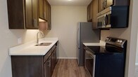 The Twain's 1-bedroom, 1-bathroom floor plan is designed for functional, comfortable living across a single level. The galley-style kitchen is a standout feature, showcasing rich dark-walnut cabinetry with modern hardware, crisp white countertops, hardwood-style flooring, and a full suite of stainless steel appliances including a refrigerator, electric range, built-in microwave, and dishwasher. Ample upper and lower cabinet storage lines both sides of the kitchen, maximizing utility in the space.

The kitchen flows directly into the living area, which is carpeted and bathed in natural light from a large window overlooking lush greenery. Crown molding accents the living room ceiling, adding a refined touch to the overall design. A wood-paneled entry door with an intercom system provides a secure and welcoming entrance.

The bedroom is generously sized with plush carpet, a large window for natural light, and includes access to a walk-in closet as well as an additional closet, providing substantial storage. Baseboard heating is present throughout, ensuring year-round comfort. The neutral, warm color palette throughout the residence creates a clean and inviting atmosphere suited to a variety of personal styles.