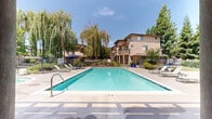 Located at 1659 Drew Ave in Davis, California, this well-appointed outdoor community amenity space offers a comprehensive array of recreational features designed for relaxed, open-air living. At the heart of the space is a generously sized swimming pool with crystal-clear water, accessible via stainless steel entry rails and surrounded by a broad concrete sun deck furnished with multiple lounge chairs — ideal for sunbathing under the California sky. Adjacent to the pool, a spa provides an additional relaxation option. A covered pergola area anchors one side of the deck, offering a shaded retreat furnished with seating and umbrella-topped tables for outdoor dining and socializing. On the opposite end of the complex, a dedicated outdoor lounge area features a built-in BBQ station and comfortable seating surrounded by vibrant blue decorative planters and manicured landscaping. A well-maintained grass lawn extends through part of the grounds, complemented by recreational amenities including a ping-pong table. The entire area is enclosed by an iron perimeter fence and framed by mature weeping willows and towering shade trees, creating a private, park-like atmosphere. Multi-story residential buildings line the perimeter, lending a cohesive, community-centered character to the space.