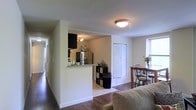 This single-floor residence offers a thoughtfully arranged open-concept layout that seamlessly connects the living, dining, and kitchen areas. The living space features rich, dark hardwood flooring that contrasts elegantly with the clean white walls, creating a warm yet modern ambiance. The kitchen is partially separated by a half-wall partition, lending a sense of definition to the space while maintaining an open feel. It is equipped with a stainless steel refrigerator and features a granite-style countertop that serves as a convenient breakfast bar or prep area. Dark cabinetry overhead adds a contemporary touch to the kitchen's design. Adjacent to the kitchen, the dining area accommodates a wooden dining table with matching chairs, positioned near a window that invites natural light into the space. A set of bi-fold closet doors provides practical storage access nearby. Throughout the residence, flush ceiling lighting ensures consistent illumination. A hallway visible from the main living area suggests additional rooms beyond, extending the functional footprint of this well-organized, single-level home.