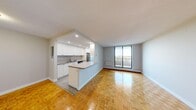 This well-organized two-bedroom, one-bathroom unit is laid out entirely on a single floor, making efficient use of its space throughout. The combined kitchen and living area forms the heart of the unit, where warm parquet flooring extends across the open-concept space. The kitchen is fitted with white cabinetry, stainless steel appliances, a peninsula with a neutral-toned countertop, and tile flooring, clearly delineating it from the living area. Recessed lighting illuminates the kitchen, while a large floor-to-ceiling sliding door in the living room fills the space with natural light and provides access to outdoor views.

Both bedrooms continue the parquet flooring and feature windows with exterior views, baseboard heating units, and neutral grey walls. The primary bedroom includes a set of sliding closet doors offering generous storage. The secondary bedroom is similarly proportioned and well-lit.

A hallway connects the living areas to the bedrooms and the full bathroom, which features a soaking tub with clean white subway tile surround and chrome fixtures. Textured ceilings are consistent throughout, contributing to the unit's cohesive, understated aesthetic.