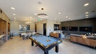 This contemporary amenity lounge is thoughtfully designed to accommodate a variety of social and recreational activities across one open, well-appointed floor. The space is anchored by a full-size billiards table positioned beneath a sleek, circular pendant light fixture, creating a natural focal point within the room. Large-format tile flooring extends throughout the main game area, lending a clean and modern aesthetic.

Adjacent to the billiards area, a dedicated games zone features a regulation ping-pong table and a game table with seating — ideal for board games and casual gatherings. Bold wall art, including a striking pop-art style canvas, adds personality and visual interest to the space.

A generously sized lounge area offers plush sectional sofas arranged around a wall-mounted flat-screen television, with patterned carpet underfoot providing a warm contrast to the tiled zones. Dark accent walls and recessed lighting create an intimate, upscale atmosphere in this seating area. Large windows allow natural light to filter in and offer views of the surrounding exterior.

A separate dining area with a long table and upholstered chairs provides a functional space for group meals or meetings, while a wet bar or kitchenette serves as a convenient refreshment station. Throughout the space, the design balances modern finishes, ambient lighting, and curated décor to create an inviting, versatile environment.