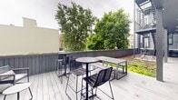 Located in the vibrant Fishtown neighborhood of Philadelphia, this contemporary outdoor amenity space at 1221 N Fourth St showcases thoughtful modern design across multiple al fresco areas. The property features a striking green roof garden, where low-growing, lush ground cover plantings create a living landscape bordered by a sleek dark gray vertical-slat perimeter fence. Gravel pathways complement the greenery, while a wooden deck area provides space for outdoor dining and relaxation with modern bench-style seating.

A welcoming courtyard deck anchors the ground level, clad in light gray composite decking and framed by the building's contemporary horizontal siding in muted sage and charcoal tones. Industrial-style metal staircases connect multiple levels, adding an architectural edge to the space. Recessed lighting under the upper balcony overhang ensures the courtyard remains functional in the evening hours. Small bistro table-and-chair sets are positioned throughout, creating intimate gathering spots.

The upper terrace offers an elevated outdoor retreat with high-top bar tables and woven rattan-style chairs, all set against a canopy of mature deciduous trees that provide natural shade and a verdant urban backdrop. The overall aesthetic is clean, modern, and community-oriented, designed to foster outdoor connection in the heart of Philadelphia.