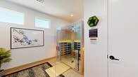 This single-floor wellness hallway is a thoughtfully designed space that blends modern aesthetics with functional amenities. The centerpiece is a glass-enclosed infrared sauna constructed with warm natural wood paneling on the interior walls, ceiling, and built-in bench seating. The sauna features chromotherapy lighting that emits vivid green and amber hues, enhancing the wellness ambiance. Full-length tempered glass panels with polished stainless steel hardware provide a sleek, transparent enclosure that allows the sauna's warm glow to illuminate the surrounding space.

The hallway itself is finished with light hardwood flooring, crisp white walls, and recessed ceiling lighting that contributes to a clean, contemporary feel. A high horizontal window admits natural light, brightening the space. A bold black-and-white speckled area rug anchors the floor, providing a striking contrast to the light tones throughout.

Decorative elements include a wall-mounted electric fireplace display flanked by a live moss shelf installation and a hexagonal moss wall accent, adding organic texture. A large framed botanical print and a potted palm introduce natural elements. Two minimalist white doors with black hardware provide access to adjoining spaces, completing this wellness-focused, design-forward environment.