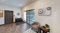 This single-floor commercial office space offers a clean, contemporary design with an open and functional layout. The interior is finished with warm hardwood-style flooring throughout, crisp white walls, and recessed lighting that keeps the space bright and welcoming.

Upon entry, a glazed front door with frosted branding graphics opens into a reception and waiting area furnished with a cushioned barrel chair, a side table with greenery, and a patterned area rug. Large sliding glass panels flood the entry with natural light and provide views of the parking area outside. Decorative wall art adds a polished touch to the space.

The main area transitions seamlessly into a refreshment and business services station along one wall, featuring dark cabinetry with a light countertop, a coffee maker, a beverage cooler stocked with drinks, and a multifunction printer on a rolling cart. A rustic wood-topped high bar table with industrial metal stools serves as a casual workspace or meeting spot, equipped with monitor mounts for convenient use.

Large interior windows look into an adjacent fitness room visible in the background, enhancing the sense of openness. A secondary door leads to a restroom corridor, indicated by signage. The overall ambiance is modern and professional, well-suited for a leasing office or small business environment.