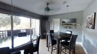 This single-floor space centers around a well-appointed dining area that combines modern and industrial design elements. The room is finished in cool gray tones on the walls, complemented by light wood-style flooring that adds warmth to the overall aesthetic. A standout feature is the ceiling fan with a distinctive teal glass globe accent, paired with a sleek pendant light, both adding character to the space.

The dining area accommodates two separate dark-toned tables with matching black leather chairs, offering flexible seating arrangements suitable for gatherings of various sizes. Large sliding glass doors along one wall flood the room with natural light and provide direct access to a balcony, where mature trees are visible, creating a pleasant outdoor connection and sense of privacy.

Decorative touches include a large photographic canvas depicting an open highway framed by snow-capped mountains, adding a dramatic focal point to the room. A louvered interior door and a motivational wall sign contribute additional visual interest. The textured ceiling and neutral palette tie the space together, creating a functional yet visually engaging environment.
