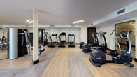 This fitness center is thoughtfully designed to accommodate a wide range of exercise routines across multiple distinct zones. The space features a clean, modern aesthetic with a neutral color palette of whites and warm beiges, accented by dark wood baseboards and black-framed glass doors that add a polished, contemporary feel.

The cardio zone is outfitted with multiple treadmills, elliptical machines, and a stair climber, all arranged on light wood-look flooring that lends a bright, open atmosphere. A dedicated cycling area features a Peloton bike positioned beneath a wall-mounted flat-screen television, set on its own wood-toned platform. A second flat-screen TV serves the strength training area, which includes a cable machine, a multi-use power rack, adjustable benches, and a full dumbbell rack with weights neatly organized on tiered shelving. Floor-to-ceiling mirrors in this zone enhance the sense of space while supporting proper form during workouts.

The flooring transitions between rubberized material in the strength area and wood-look planks elsewhere, offering both comfort and durability. Recessed and linear lighting throughout ensures consistent illumination, while a glass door provides a connection to the outdoors, contributing to an energizing and welcoming environment.