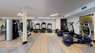 This fitness center is thoughtfully designed to accommodate a wide range of exercise routines across multiple distinct zones. The space features a clean, modern aesthetic with a neutral color palette of whites and warm beiges, accented by dark wood baseboards and black-framed glass doors that add a polished, contemporary feel.

The cardio zone is outfitted with multiple treadmills, elliptical machines, and a stair climber, all arranged on light wood-look flooring that lends a bright, open atmosphere. A dedicated cycling area features a Peloton bike positioned beneath a wall-mounted flat-screen television, set on its own wood-toned platform. A second flat-screen TV serves the strength training area, which includes a cable machine, a multi-use power rack, adjustable benches, and a full dumbbell rack with weights neatly organized on tiered shelving. Floor-to-ceiling mirrors in this zone enhance the sense of space while supporting proper form during workouts.

The flooring transitions between rubberized material in the strength area and wood-look planks elsewhere, offering both comfort and durability. Recessed and linear lighting throughout ensures consistent illumination, while a glass door provides a connection to the outdoors, contributing to an energizing and welcoming environment.