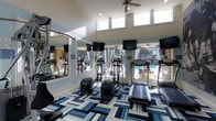 This fitness center is designed to support a comprehensive cardio-focused workout experience within a bright, open single-floor layout. The space features soaring ceilings with clerestory windows positioned near the roofline, allowing abundant natural light to fill the room alongside large floor-to-ceiling windows that frame views of an outdoor pool area. A ceiling fan overhead aids in air circulation, contributing to a comfortable workout environment.

The room is outfitted with a selection of cardio equipment, including treadmills, elliptical trainers, and stationary bikes, each accompanied by wall-mounted flat-screen televisions for entertainment during workouts. Full-length mirrors line one wall, enhancing the sense of space and allowing users to monitor their form. Stone accent wall detailing adds a decorative architectural touch, contrasting with the clean white walls and blue accent stripes that define the room's color palette.

Underfoot, a bold geometric-patterned carpet in shades of navy, sky blue, and white adds a dynamic visual element to the space. The overall ambiance is energizing and modern, with ample natural light and outdoor views creating an inviting atmosphere for fitness activity.