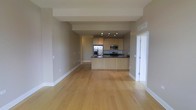 This single-floor space showcases a thoughtfully designed open-concept layout that effortlessly blends the living area and kitchen into one cohesive environment. The expansive main living area is anchored by warm, light-toned hardwood flooring that runs continuously throughout, lending a clean and inviting feel to the space.

The kitchen is positioned toward the rear and features light maple cabinetry that offers ample storage. A dark-toned countertop provides a striking contrast against the cabinetry, while the peninsula extends the counter space and creates a natural visual boundary between the kitchen and living areas. Stainless steel appliances, including a refrigerator, microwave, and range, are neatly integrated into the kitchen design, adding a modern and functional touch.

Track lighting installed above the kitchen area ensures targeted illumination for meal preparation, while the overall neutral color palette — consisting of soft warm-gray tones on the walls and crisp white trim — creates a bright and airy ambiance. A door on the right side of the space leads to an additional room or exterior access, contributing to the functional flow of the layout. The combination of open space, modern finishes, and a cohesive design makes this a highly livable and versatile environment.