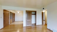 This single-story space is characterized by its warm, light-toned hardwood flooring that extends throughout the visible areas, contributing to a cohesive and inviting atmosphere. The interior is finished with neutral white walls that create a bright, open feel, complemented by standard-height ceilings with ceiling fan fixtures providing both air circulation and ambient lighting.

A central feature of the main area is a set of louvered bi-fold closet doors with warm wood tones, offering built-in storage with an open shelving compartment above. The layout reveals multiple connected rooms, with doorways leading to additional spaces, including what appears to be a secondary room visible in the background, also equipped with a ceiling fan and its own louvered closet doors. A hallway is visible to the right, hinting at further rooms beyond.

HVAC vents are integrated into the upper walls, ensuring climate control throughout. The overall design reflects a straightforward, functional aesthetic with consistent wood-toned accents across doors and trim, giving the space a unified, traditional character suitable for a variety of uses.