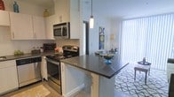 This single-floor residence offers a thoughtfully designed open-concept layout that seamlessly connects the kitchen and living areas, creating a bright and welcoming atmosphere. The kitchen is a standout feature, showcasing crisp white cabinetry that extends to the upper walls, providing ample storage space. Dark granite-style countertops offer a bold contrast against the white finishes, while stainless steel appliances — including a range, microwave, and additional built-in units — lend a sleek, contemporary look. A peninsula-style counter extends into the space, offering additional prep area and a natural visual divider between the kitchen and the living area. Pendant lighting above the counter adds a warm, focused glow that enhances the modern aesthetic. The adjoining living area is bathed in natural light streaming through floor-to-ceiling vertical blinds, creating an open and airy feel. Light hardwood-style flooring runs throughout, tying the spaces together cohesively. The overall ambiance is clean, modern, and inviting, with a neutral color palette accented by thoughtful decor, making it a comfortable and functional living environment.