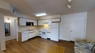 This single-floor space offers a practical and well-organized layout designed for comfortable everyday living. The kitchen is a standout feature, showcasing an L-shaped configuration with ample white cabinetry accented by warm wood-tone trim, providing generous storage throughout. The countertops offer plenty of workspace, and the kitchen is equipped with a full suite of white appliances, including a refrigerator, electric range with overhead ventilation, dishwasher, and a built-in microwave. A stainless steel sink is conveniently positioned beneath overhead cabinets, maximizing functionality. The flooring throughout features rich, dark hardwood-style planks that add warmth and a cohesive aesthetic to the space. Overhead lighting, including a fluorescent fixture above the kitchen work area and a track light in the adjoining space, ensures the area is well-lit. A door off the kitchen provides convenient access, likely leading to a utility area or exterior. The overall ambiance is clean, neutral, and move-in ready, with a straightforward design that emphasizes practicality and ease of use.