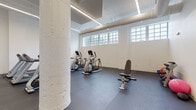 This fitness center occupies a single open floor with a clean, industrial aesthetic defined by white-painted walls, exposed cylindrical support columns, and visible overhead ductwork. The dark rubber flooring provides a durable, functional surface throughout the space, while a row of clerestory windows along one wall allows natural light to filter in, enhancing the bright, energetic atmosphere. Linear LED lighting fixtures are recessed and surface-mounted along the ceiling, ensuring consistent illumination across the entire area.

The cardio zone is well-stocked with Matrix-brand treadmills, elliptical trainers, and both upright and recumbent stationary bikes, arranged in an orderly layout that maximizes the available floor space. A motivational wall mural reading "No Goal Was Ever Met Without a Little Sweat" serves as a focal point, adding character to the otherwise minimalist interior. The strength and functional training area features a multi-functional cable machine, an adjustable weight bench, a dumbbell rack, and a stability ball, offering a comprehensive range of workout options. Wall-mounted flat-screen televisions are positioned for easy viewing from the cardio equipment. The overall layout is open and uncluttered, promoting ease of movement throughout.