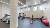 This fitness center occupies a single open floor with a clean, industrial aesthetic defined by white-painted walls, exposed cylindrical support columns, and visible overhead ductwork. The dark rubber flooring provides a durable, functional surface throughout the space, while a row of clerestory windows along one wall allows natural light to filter in, enhancing the bright, energetic atmosphere. Linear LED lighting fixtures are recessed and surface-mounted along the ceiling, ensuring consistent illumination across the entire area.

The cardio zone is well-stocked with Matrix-brand treadmills, elliptical trainers, and both upright and recumbent stationary bikes, arranged in an orderly layout that maximizes the available floor space. A motivational wall mural reading "No Goal Was Ever Met Without a Little Sweat" serves as a focal point, adding character to the otherwise minimalist interior. The strength and functional training area features a multi-functional cable machine, an adjustable weight bench, a dumbbell rack, and a stability ball, offering a comprehensive range of workout options. Wall-mounted flat-screen televisions are positioned for easy viewing from the cardio equipment. The overall layout is open and uncluttered, promoting ease of movement throughout.