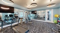 This 640 square foot fitness center is a testament to modern design and functionality. The space is characterized by its light blue walls, which create a refreshing and energizing atmosphere. Large windows allow ample natural light to flood the room, enhancing the open feel of the space.

The facility is equipped with a comprehensive range of exercise equipment. The cardio area features treadmills, elliptical machines, and stationary bikes, while the strength training section includes a variety of weight machines and free weights. A dedicated area for floor exercises is also present, complete with exercise balls and mats.

The interior design is both practical and aesthetically pleasing. Circular LED light fixtures hang from the ceiling, providing even illumination throughout. Mirrored walls not only make the space appear larger but also serve a functional purpose for those monitoring their form during workouts.

The flooring is a distinctive gray pattern, which not only adds visual interest but is also likely chosen for its durability and ease of maintenance. White-framed glass doors separate different areas of the gym, maintaining an open concept while allowing for some division of space.

Safety features are evident, with an emergency exit clearly marked. The overall layout is spacious, allowing for comfortable movement between equipment. This well-designed fitness center offers a inviting and efficient environment for various workout routines.
