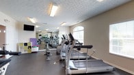 Located at Parkway Village in Grove City, Ohio, this single-floor fitness center is a well-equipped amenity space designed to support an active lifestyle. The room features a row of modern cardio machines, including treadmills and ellipticals, arranged along one side to maximize floor space and usability. A full wall of mirrors runs the length of the room, visually expanding the space and allowing members to monitor their form during workouts. Natural light filters in through an elegant arched window, brightening the interior and creating an open, welcoming atmosphere. The flooring consists of durable rubber matting, providing both safety and comfort underfoot. Overhead, patterned ceiling tiles add a subtle decorative touch while flush-mounted fluorescent fixtures ensure consistent, even lighting throughout. A wall-mounted flat-screen television is positioned for easy viewing from the cardio area. Along the opposite wall, colorful storage elements and additional fitness accessories suggest a dedicated zone for stretching or group exercise. The neutral, beige-toned walls contribute to a clean and focused environment. Overall, the fitness center is thoughtfully laid out to accommodate a variety of workout routines in a comfortable and functional community setting.