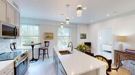 Located at 181 North Main Street in Suffolk, Virginia, this thoughtfully designed one-bedroom, one-bathroom residence offers a comfortable and efficient single-floor layout. The open-concept kitchen serves as the heart of the home, showcasing a large center island with a white quartz countertop and undermount sink, elegant pendant lighting, and taupe shaker-style cabinetry. A herringbone tile backsplash complements the full suite of stainless steel appliances, including a French door refrigerator, electric range, and over-the-range microwave. Multiple windows flood the kitchen and adjoining dining area with natural light, framing views of lush greenery outside. The bedroom is a serene retreat, featuring a ceiling fan, a walk-in closet, and large windows that continue the home's bright and airy feel. Light-toned flooring flows consistently throughout the space, enhancing the sense of openness. The bathroom is clean and functional, finished with white cabinetry, a single vanity with brushed nickel hardware, and a large mirror. Crown molding and crisp white walls add refined architectural detail throughout, creating a cohesive and polished aesthetic across every room.