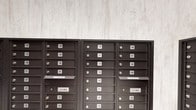 This mailroom serves as a centralized mail distribution hub within a larger residential building, accommodating a substantial number of units as evidenced by the extensive numbering system spanning from the low 100s through at least the 280s. The space is organized across multiple wall sections, each lined with dark bronze-finished, individually numbered mailboxes arranged in neat vertical and horizontal rows. Integrated U.S. Mail slots are positioned throughout the banks, allowing postal carriers convenient and secure delivery access.

The design of the mailroom balances functional necessity with thoughtful aesthetics. Two distinct wall treatments define the space: one area features light-toned, textured stone-look wall panels that lend a clean, contemporary backdrop to the dark metal mailboxes, while another section showcases a striking reclaimed wood accent wall with varied plank tones ranging from warm amber to cool grey. Light-toned tile flooring runs throughout, contributing to a bright, open feel. A dark paneled door is visible in the background, providing a refined transition point into adjacent areas of the building. The overall ambiance is modern yet welcoming, blending practical utility with design-conscious finishes.