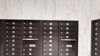 This mailroom serves as a centralized mail distribution hub within a larger residential building, accommodating a substantial number of units as evidenced by the extensive numbering system spanning from the low 100s through at least the 280s. The space is organized across multiple wall sections, each lined with dark bronze-finished, individually numbered mailboxes arranged in neat vertical and horizontal rows. Integrated U.S. Mail slots are positioned throughout the banks, allowing postal carriers convenient and secure delivery access.

The design of the mailroom balances functional necessity with thoughtful aesthetics. Two distinct wall treatments define the space: one area features light-toned, textured stone-look wall panels that lend a clean, contemporary backdrop to the dark metal mailboxes, while another section showcases a striking reclaimed wood accent wall with varied plank tones ranging from warm amber to cool grey. Light-toned tile flooring runs throughout, contributing to a bright, open feel. A dark paneled door is visible in the background, providing a refined transition point into adjacent areas of the building. The overall ambiance is modern yet welcoming, blending practical utility with design-conscious finishes.