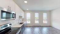 This contemporary single-floor unit showcases a clean, modern design throughout. The open-concept layout seamlessly connects the kitchen and living area, creating a bright and airy atmosphere enhanced by large double-hung windows that flood the space with natural light and offer views of the surrounding neighborhood. Wood-look plank flooring runs throughout, tying the spaces together cohesively.

The kitchen is a standout feature, outfitted with a two-tone cabinetry scheme pairing crisp white upper cabinets with wood-grain lower cabinets. A suite of stainless steel appliances includes a refrigerator, electric range, built-in microwave, and dishwasher, all complemented by light quartz countertops and a stainless steel sink. Recessed and flush-panel ceiling lighting provides well-distributed illumination.

The bathroom continues the unit's modern aesthetic with white cabinetry, a sleek white vanity countertop, a large mirror, and wood-look tile flooring, creating a clean and functional space. A dedicated utility area houses a stacked washer and dryer unit alongside built-in wire shelving, offering convenient in-unit laundry and additional storage. Overall, the unit presents a polished, move-in-ready living environment with thoughtful finishes throughout.