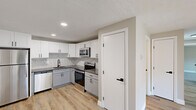 Located at 556 Mill St in Worcester, MA, this 2-bedroom, 1-bathroom unit offers a well-designed single-floor layout with modern finishes throughout. The kitchen is a standout feature, showcasing white and gray shaker-style cabinetry, a herringbone tile backsplash, dark countertops, and a full suite of stainless steel appliances including a refrigerator, range, over-the-range microwave, and dishwasher. Light-toned luxury vinyl plank flooring runs through the kitchen and living areas, lending a clean, contemporary feel. The combined kitchen and living room area provides a practical, open flow for everyday living. A sliding glass door in the living room fills the space with natural light and opens to a private balcony with outdoor views. Both bedrooms are finished with soft gray carpet, neutral warm-toned walls, and baseboard heating. The primary bedroom offers a comfortable, well-proportioned layout with a closet. The bathroom features white subway tile surrounding the tub-shower combo, a stone-top vanity, warm wood-toned LVP flooring, and matte black hardware and fixtures, creating a cohesive, modern aesthetic throughout.
