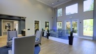 This single-floor space presents a sleek and contemporary lobby area designed to impress visitors from the moment they arrive. The architecture is defined by dramatically high ceilings that amplify the sense of openness, complemented by a wall of tall, expansive windows that flood the interior with abundant natural light and offer views of the surrounding lush greenery.

The flooring throughout is a warm, light-toned hardwood that anchors the space with a clean, modern aesthetic. A welcoming seating arrangement, featuring a stylish gray lounge chair accented with a colorful throw pillow and set upon a rich blue area rug, adds a touch of comfort and color to the reception area.

A reception desk, identifiable by branded signage, serves as the functional centerpiece of the lobby and is tastefully integrated into the overall design. Potted plants in sleek black planters are strategically placed throughout, introducing natural elements that soften the modern lines of the space.

The neutral gray wall palette paired with white trim creates a crisp, professional backdrop, while subtle blue accent lighting near the reception area adds a contemporary and inviting ambiance. An adjacent office or administrative area is visible through a doorway, offering a glimpse of the functional spaces beyond the main lobby.