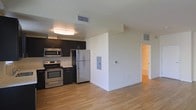 This single-floor space showcases a thoughtfully designed open-concept layout that seamlessly connects the kitchen and living area. Light hardwood flooring runs throughout, lending warmth and continuity to the space.

The kitchen is outfitted with dark espresso cabinetry that provides a striking contrast against the light-colored countertops and walls. A full suite of stainless steel appliances includes a refrigerator, range with oven, microwave, and dishwasher, offering a well-equipped cooking environment. A window above the sink brings in additional natural light, while recessed ceiling lighting further illuminates the workspace.

The open living area is bright and airy, benefiting from a large sliding glass door that leads to a private balcony with a metal railing. This feature allows natural light to flood the interior and provides a direct indoor-outdoor connection. The neutral wall tones and clean architectural lines create a contemporary, understated ambiance throughout.

A hallway visible from the main living area leads to additional rooms, suggesting a practical and well-organized floor plan. Overall, the space presents a clean, modern aesthetic with functional, quality finishes.