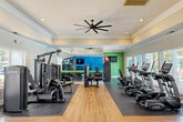 This fitness center in Birmingham, Alabama offers a thoughtfully designed, single-level workout space with a generous open floor plan. The interior features light hardwood-style flooring complemented by rubber mat zones that define the strength and cardio areas, providing both functionality and visual organization throughout the space.

The cardio section is lined along a wall of large windows that flood the room with natural light and offer views of the outdoor surroundings, including what appears to be a pool area. This section is equipped with multiple True-brand treadmills, elliptical trainers, and a stationary cycle, each outfitted with individual display screens. A large ceiling fan anchors the tray ceiling above, enhancing airflow and comfort during workouts.

The strength training area features a selection of True-brand cable and resistance machines, a dedicated free weights rack with dumbbells, a rowing machine, and a wall-mounted flat-screen television set against a bold blue accent wall. Full-length mirrors along one wall visually expand the space and assist with form during exercises.

Crown molding, recessed lighting, and a tray ceiling with decorative coffered detailing contribute to a polished, upscale aesthetic. A glass-paneled door provides direct exterior access. The overall ambiance is bright, clean, and motivating, making this fitness center a well-appointed facility for a comprehensive workout experience.