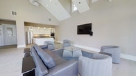 Located at 193 Tenby Chase Drive in Delran, New Jersey, this single-floor space is designed with an open-concept layout that promotes a sense of airiness and flow. The most striking architectural feature is the dramatic vaulted ceiling, which rises to impressive heights and is complemented by strategically placed skylights and high windows that flood the interior with abundant natural light.

The main living area is furnished with plush, contemporary seating including a large leather sofa and coordinating accent chairs in soft blue-gray tones, all arranged around a glass coffee table atop a blue area rug. A wall-mounted flat-screen television anchors one side of the room, making this an ideal space for gatherings and entertainment.

Seamlessly connected to the living area is a modern kitchen outfitted with crisp white cabinetry, stainless steel appliances including a refrigerator and double wall ovens, and a spacious island with pendant lighting above. The clean, neutral color palette of warm white walls and light flooring throughout creates a cohesive and inviting atmosphere. This well-appointed space is well-suited for community events, social gatherings, or multi-purpose use.