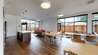 Located at 9747 NE Glisan St in Portland, Oregon, this single-floor communal space is designed with a contemporary, open-plan layout that seamlessly integrates multiple functional zones. The interior is defined by rich, dark hardwood flooring that runs throughout, crisp white walls, and an abundance of natural light delivered through expansive black-framed floor-to-ceiling windows that frame views of an outdoor patio and surrounding greenery.

The lounge area features comfortable seating arrangements, including bold rust-orange sofas and armchairs paired with light wood accent tables, creating a warm and inviting gathering environment. Large sliding glass doors connect this area directly to an outdoor patio space, further enhancing the sense of openness. Overhead, oversized drum pendant lights provide consistent, ambient illumination.

A flexible co-working and dining zone occupies a generous portion of the floor plan, furnished with movable tables and patterned chairs arranged to accommodate both individual use and group activity. This area benefits from multi-directional natural light through wraparound windows.

The kitchen area is equipped with dark espresso cabinetry, a stainless steel refrigerator, a built-in microwave, and a sleek white-topped island, providing a practical and polished food preparation space. A smaller meeting nook with interior windows offering views into the main area rounds out the layout, making this a well-appointed, versatile community amenity space.