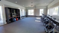 This fitness center offers a clean, well-organized workout environment designed to accommodate a variety of exercise routines. The space features a neutral color palette with gray-toned walls accented by a dark wainscoting band, complemented by gray carpet flooring that runs throughout the room. Recessed lighting and a central ceiling fan with a light fixture ensure the space remains bright and comfortable during workouts. Large windows along two walls flood the interior with abundant natural light while providing views of the surrounding greenery outdoors. On one side of the room, a dark shelving unit stores an assortment of colorful medicine balls and exercise balls of varying sizes, keeping equipment neatly organized and accessible. Upright cardio machines, including treadmills and ellipticals, are lined along the opposite wall, offering ample workout stations. The open central floor area provides generous space for stretching, floor exercises, or group fitness activities. The overall ambiance is functional yet inviting, with a well-maintained layout that balances equipment storage and open floor space to create a versatile and practical fitness environment.