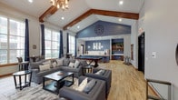 This rentable social lounge is a thoughtfully designed communal space that blends comfort, style, and functionality. The room is anchored by soaring vaulted ceilings accented with rich, dark-stained exposed wood beams, creating a warm yet airy atmosphere throughout. Light hardwood flooring runs the length of the space, complementing the neutral gray and blue color palette that gives the lounge a cohesive, modern aesthetic.

The main living area features generously sized gray upholstered sofas and armchairs arranged around coffee tables, providing ample seating for socializing. A classic fireplace with a white mantle serves as a focal point on one wall, topped by a mounted flat-screen television flanked by wall sconces. Tall, multi-pane windows dressed with navy drape curtains line the walls, bathing the interior in natural light and offering views of the surrounding greenery.

A centrally placed pool table adds a recreational element, while high-top bistro tables positioned near the windows offer intimate spots for conversation. The back of the lounge transitions into a well-equipped kitchenette, complete with warm wood cabinetry, a light stone countertop, a stainless steel refrigerator, a sink, and open shelving adorned with greenery. A bold blue accent wall ties the kitchen area together, adding depth and visual interest. This versatile lounge is well-suited for private events, community gatherings, or casual entertaining.