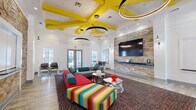 This single-floor space makes a strong visual statement through its bold, contemporary design. The entrance greets visitors with glass double doors, transom windows, and light wood-style flooring that extends throughout. A branded welcome mat and a small refreshment station establish a welcoming, communal atmosphere from the outset.

The main lounge area features soaring ceilings accented by a distinctive grid of vivid yellow structural beams, complemented by multiple large circular pendant light fixtures that provide dramatic, layered illumination. A full-height shimmering curtain partition serves as an artistic room divider, while a pair of chartreuse accent chairs flanks a sculptural white side table nearby.

The lounge's focal point is a floor-to-ceiling stacked stone accent wall housing a large flat-screen television and a floating shelf. Surrounding this anchor are bold, colorful sectional sofas, including a vivid red-and-teal configuration and a multicolored striped ottoman, arranged over a rich, dark area rug. Sculptural metallic coffee tables add further visual interest.

A high-top seating area with bar-height chairs provides a functional workspace or social zone near glass doors that open to an outdoor area. Recessed lighting and wall sconces supplement the pendant fixtures throughout, ensuring the space is well-lit and energetic at all times.