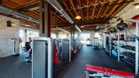 This expansive fitness facility masterfully blends industrial aesthetics with a fully equipped workout environment. The space is defined by its striking architectural character: exposed wood-beam and rafter ceilings soar overhead, complemented by visible HVAC ductwork and warm pendant lighting that together create an authentic industrial ambiance. The perimeter walls feature raw, whitewashed brick, adding texture and a sense of history to the interior.

Large steel-framed windows line the walls, flooding the facility with abundant natural light and offering open views of the surrounding outdoor landscape. The floor is finished with durable rubber and interlocking tile surfaces, providing both safety and comfort for workouts.

The main exercise floor is generously sized and organized into distinct zones. A robust selection of strength training machines, finished in silver and red, are arranged in rows along the central space, offering a wide range of resistance equipment. A dedicated cardio zone features multiple treadmills, ellipticals, cycles, and rowing machines positioned to take full advantage of the window views. A free-weight and functional training area rounds out the offerings.

Near the entrance, an industrial shelving unit displays rolled towels alongside decorative "GYM" lettering, reinforcing the facility's identity. The overall atmosphere is energetic yet refined, combining purposeful design with a welcoming, well-lit environment.