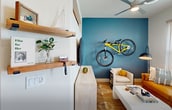 This compact yet stylish 440 square foot apartment maximizes space through thoughtful design. The open-concept layout seamlessly integrates living, sleeping, and kitchen areas. The living room boasts a striking blue accent wall, complemented by a yellow sofa and white coffee table. A unique feature is the wall-mounted bicycle, adding a functional and artistic touch.

The kitchen is a model of efficiency, with white cabinetry, stainless steel appliances, and a herringbone backsplash. It includes a full-size refrigerator, range, and built-in microwave. Open shelving adds both storage and display space.

The sleeping area, while not fully enclosed, is defined by a blue accent wall and features a comfortable bed with cheerful yellow accents. A transom window above allows natural light to filter through.

The bathroom, though compact, offers a full-size tub with a curved shower rod and a modern vanity. Throughout the apartment, hardwood floors add warmth and continuity. Clever storage solutions, including built-in closets and shelving, maximize the available space. Large windows flood the space with natural light, creating an airy atmosphere despite the limited square footage.