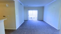 This single-floor space features an open living area finished with wall-to-wall carpet in a neutral, speckled tone that adds texture underfoot while contributing to a quiet, comfortable atmosphere. The room is defined by clean white walls accented with crown molding along the ceiling line, lending a polished, cohesive look to the interior. A sliding glass door positioned along the far wall is fitted with vertical blinds, allowing for adjustable natural light and providing access to an outdoor area beyond. The overall layout is straightforward and open, offering a blank canvas well-suited to a variety of furniture arrangements. Recessed ceiling fixtures provide overhead lighting, while standard white baseboards frame the perimeter of the room cleanly. A light switch panel is visible near the entryway, and a ceiling vent indicates a central HVAC system. The neutral color palette throughout — from the flooring to the walls and trim — creates a bright, airy feel. The space is practical and uncluttered, with a layout that maximizes usable floor area.