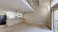 Located at 200 Carmen Ave in Carrboro, NC, this two-story residence offers a distinctive loft-style layout that maximizes vertical space and creates a dynamic, open feel throughout. The main level centers on a generous living area with soaring ceilings, neutral carpet flooring, and a sliding glass door that opens directly onto a private wooden balcony, bringing in natural light and outdoor views of surrounding greenery. An open-concept kitchen adjoins the living space and features white cabinetry, gray laminate countertops, a peninsula breakfast bar, and black appliances including a refrigerator, electric range, dishwasher, and built-in microwave. A stacked washer and dryer unit is conveniently situated on the main level. Two bathrooms serve the residence: one featuring a tiled tub-shower combination with wood-look flooring, and another with a white vanity, gray countertop, and a window for natural light. A staircase leads to the upper loft level, which is open to the living area below via a white railing. This upper floor offers a spacious carpeted area with a ceiling fan, a closet, and access to an additional bathroom, making it well-suited for use as a primary bedroom or flexible living space.