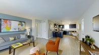 This single-floor residence offers a thoughtfully designed open-concept layout that seamlessly connects the living, dining, and kitchen areas into one cohesive and functional space. The living area features light hardwood-style flooring that transitions into soft carpeting, anchoring a comfortable seating arrangement accented by bold orange chairs and a classic sofa. A soft blue-grey accent wall adds a touch of color and character to the space, while light grey walls throughout maintain a clean, contemporary aesthetic. The kitchen is a standout feature, showcasing deep navy cabinetry paired with stainless steel appliances, pendant lighting, and a breakfast bar with seating — ideal for casual dining or entertaining. A dedicated dining nook with a modern table and chairs sits adjacent to the kitchen, bathed in natural light from a nearby window. Recessed and track lighting complement the pendant fixtures, ensuring the space is well-lit throughout. Smooth white ceilings contribute to an open and airy atmosphere. Hallway access to additional rooms is visible from the main living area, suggesting a practical and well-organized floor plan that balances style with everyday livability.
