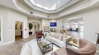 This newly renovated clubhouse, known as The Club, spans a generous single floor and offers a sophisticated, welcoming environment designed for comfort and community. The main lounge serves as the centerpiece, featuring a dramatic multi-tiered tray ceiling with a high-gloss finish, recessed lighting, and rich stone-look tile flooring accented by striking marble-patterned inlays. Elegantly furnished with plush seating, leather accent chairs, and glass-top coffee tables, this space exudes a refined yet approachable ambiance. Arched doorways frame transitions between areas, lending a classic architectural character throughout.

The dining and café area is bathed in natural light from expansive windows and glass French doors that open to outdoor views, creating an airy atmosphere ideal for gathering. A beverage station, bold abstract artwork, and a statement globe chandelier add personality and warmth to this zone. Multiple seating arrangements, from round glass tables to intimate lounge chairs, accommodate a variety of uses.

A dedicated entertainment lounge features a substantial built-in wooden media wall with integrated shelving and a large flat-screen display, flanked by solid wood benches atop marble tile flooring. The full kitchen is outfitted with crisp white cabinetry, stainless steel appliances, a subway tile backsplash, quartz countertops, and stylish pendant lighting, completing this fully functional and thoughtfully designed communal space.
