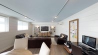 This single-floor space presents a thoughtfully designed living area that combines contemporary and industrial aesthetics. The room is characterized by a soft blue ceiling fitted with track lighting, which provides adjustable, even illumination throughout the space. The walls feature white shiplap paneling, lending a clean, textured backdrop that complements the warm hardwood-style flooring below.

The seating arrangement centers around rich, dark brown leather sofas positioned around a neutral area rug, creating a comfortable and cohesive lounge area. A wall-mounted flat-screen television serves as the focal point of the room, flanked by industrial-style open shelving units constructed from metal piping and wood, housing books and decorative items. A wooden media console sits beneath the TV, providing functional storage.

A wall-mounted mini-split HVAC unit ensures climate control, while small windows allow natural light to filter in. To the right, a cork bulletin board framed in warm wood adds a practical organizational element. The overall ambiance is relaxed yet functional, making the space well-suited for both leisure and everyday living.
