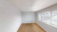 This single-story space presents a practical and straightforward layout, as seen in the overhead floor plan view, which reveals distinct zones for living, sleeping, kitchen, and bathroom functions. Throughout the carpeted rooms, warm beige flooring contrasts with light gray walls and white trim, lending a clean, neutral aesthetic. Textured ceilings are consistent across the space, adding a subtle visual element overhead.

The bedroom areas are well-proportioned, each featuring windows fitted with horizontal blinds that invite natural light while maintaining privacy. The rooms offer a blank canvas, with ample wall space and outlets positioned conveniently along the baseboards.

The kitchen is equipped with white cabinetry — both upper and lower — providing generous storage. A double-basin stainless steel sink sits beneath the upper cabinets, accompanied by laminate countertops and a white range. The layout supports efficient use of the available workspace.

The bathroom includes a full tub-shower combination with a white surround, a toilet, and a wall-mounted mirror, all finished in a clean white and neutral gray palette. Overall, the space maintains a consistent, understated design throughout, prioritizing functionality and practicality.