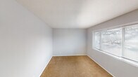 This single-story space presents a practical and straightforward layout, as seen in the overhead floor plan view, which reveals distinct zones for living, sleeping, kitchen, and bathroom functions. Throughout the carpeted rooms, warm beige flooring contrasts with light gray walls and white trim, lending a clean, neutral aesthetic. Textured ceilings are consistent across the space, adding a subtle visual element overhead.

The bedroom areas are well-proportioned, each featuring windows fitted with horizontal blinds that invite natural light while maintaining privacy. The rooms offer a blank canvas, with ample wall space and outlets positioned conveniently along the baseboards.

The kitchen is equipped with white cabinetry — both upper and lower — providing generous storage. A double-basin stainless steel sink sits beneath the upper cabinets, accompanied by laminate countertops and a white range. The layout supports efficient use of the available workspace.

The bathroom includes a full tub-shower combination with a white surround, a toilet, and a wall-mounted mirror, all finished in a clean white and neutral gray palette. Overall, the space maintains a consistent, understated design throughout, prioritizing functionality and practicality.