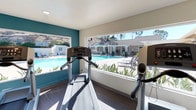 The gym at Sweetwater Springs in Spring Valley is a well-appointed, single-story fitness space designed to make workouts both functional and visually engaging. The interior is characterized by large picture windows that span much of the exterior wall, flooding the space with abundant natural light and offering unobstructed views of the adjacent outdoor pool area and surrounding hillside landscape. This seamless visual connection to the outdoors creates an open, airy atmosphere that enhances the overall workout experience.

The flooring features dark hardwood-style planks, providing a clean, modern aesthetic that contrasts nicely with the bright white walls and a bold teal accent wall that adds a pop of color and visual interest. The ceiling is smooth and well-lit with recessed lighting to supplement natural light during all hours of operation.

Cardio equipment, including a treadmill and additional exercise machines, is positioned to take full advantage of the pool and hillside views. The layout is thoughtfully organized to maximize usable floor space while maintaining an uncluttered, comfortable environment. Overall, the gym offers a bright, motivating setting with a strong emphasis on natural light and outdoor connectivity.