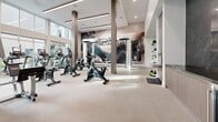 Located at 27930 Cabot Rd in Laguna Niguel, CA, this professionally designed fitness center offers a spacious, single-floor layout with a generous open floor plan. The facility is flooded with natural light through floor-to-ceiling windows that frame views of the surrounding palm-lined exterior, creating an inviting and energizing atmosphere.

The interior features soaring ceilings with sleek linear lighting and a neutral color palette of whites and warm beiges that gives the space a clean, contemporary aesthetic. A durable, light-toned rubber flooring spans the entire area, providing both functionality and comfort underfoot.

The fitness floor is organized into distinct training zones. A cardio section is outfitted with multiple stationary bikes, treadmills, elliptical machines, and cable equipment. A dedicated strength area includes a multi-station cable machine, squat racks, bench press stations, and an extensive free-weight rack with a floor-to-ceiling mirror wall. A functional training zone features ceiling-mounted TRX suspension trainers, wall-mounted ladder bars, a boxing bag, and accessory storage.

A bold, large-scale abstract mural serves as a striking focal point along one wall, adding an artistic dimension to the space. The overall design balances high performance with upscale aesthetics, making it a well-rounded and visually engaging fitness destination.